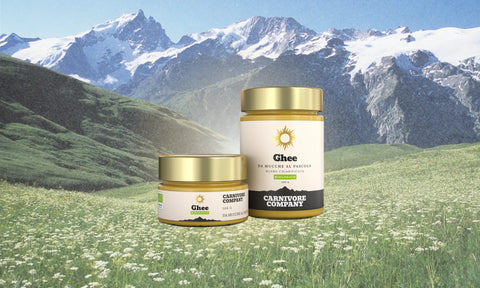 Due vasetti di Ghee biologico Carnivore Company da mucche al pascolo, burro chiarificato in formato 100g e 300g con tappo dorato, su sfondo di prato alpino fiorito e montagne innevate