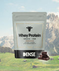 Confezione di proteine Whey grass fed al cioccolato Carnivore Company con sfondo di montagne alpine e pezzi di cioccolato.
